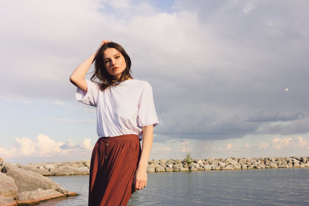 A young woman with long hair poses by a rocky lakeside, under an overcast sky.