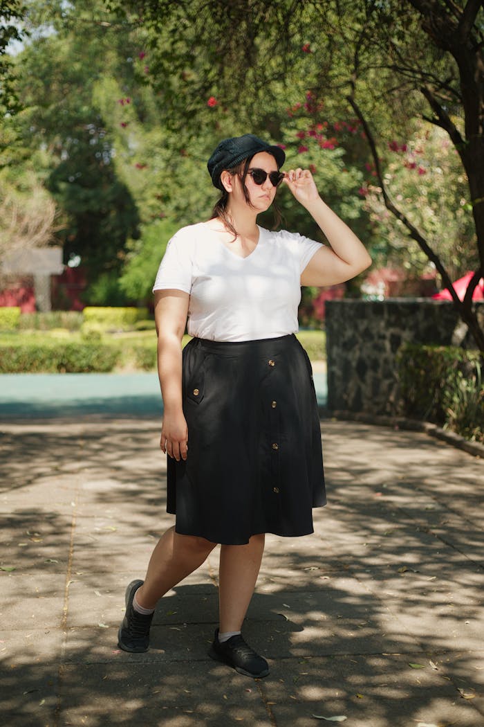 A fashionable woman in sunglasses and a skirt poses in a sunny park.