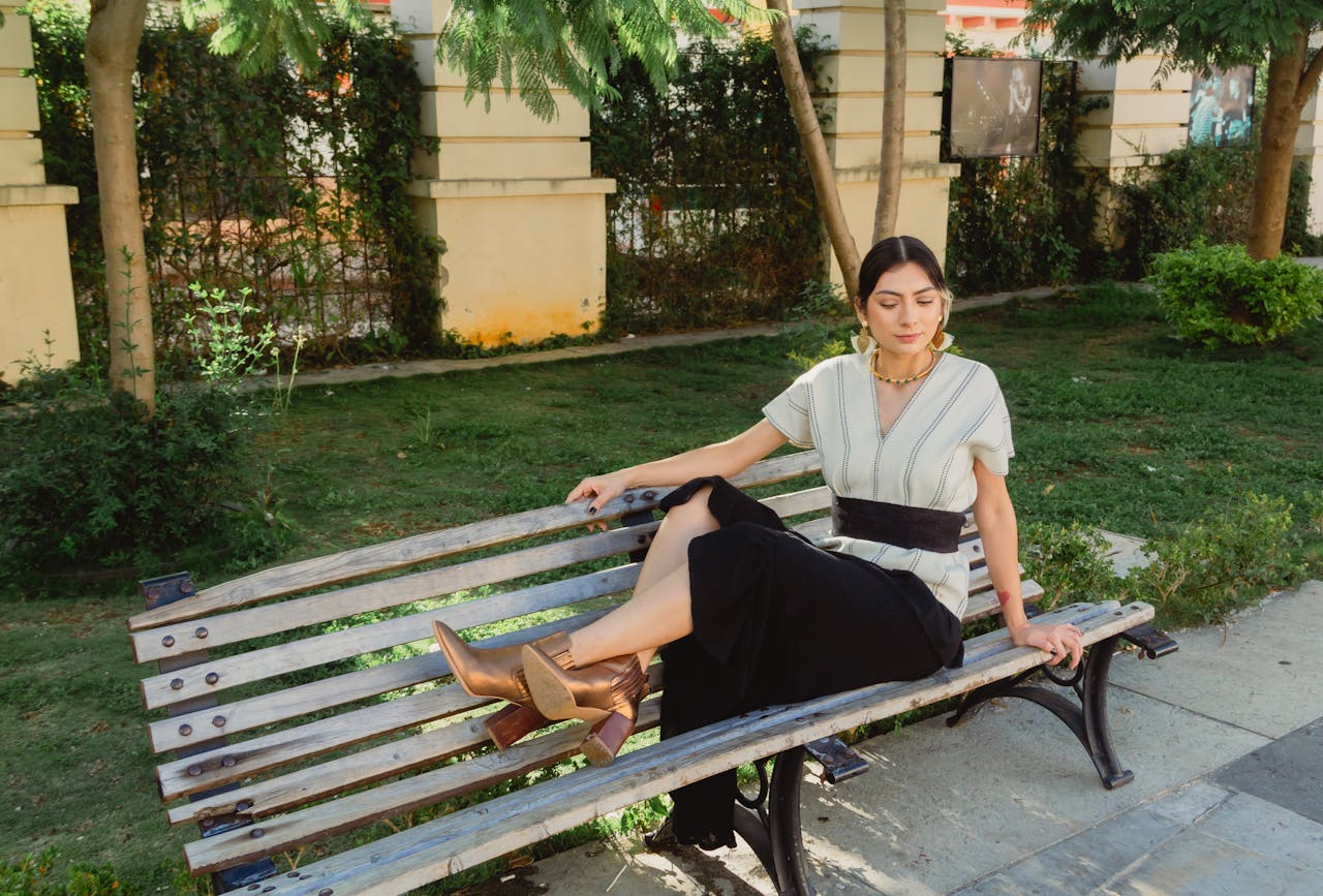 A woman sits leisurely on a park bench in a sunlit garden, surrounded by greenery.
